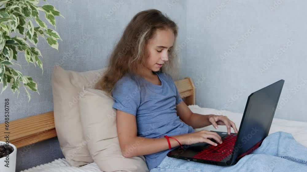 A teenage girl with an open laptop is sitting on her bed typing in her bedroom A teenage girl with an open laptop is sitting on her bed typing in her bedroom