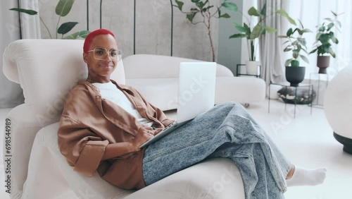 Young successful African American woman freelancer finishes typing article on laptop and smilingly looks at screen rejoicing at presence remote work sits in comfortable chair in room with house plants photo