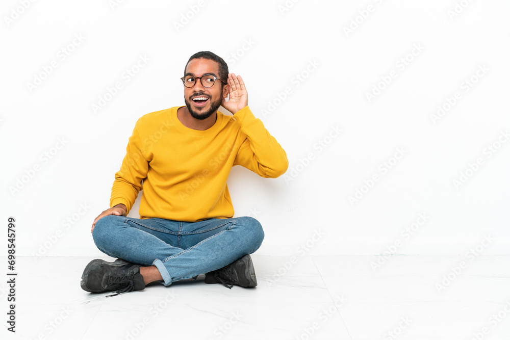 Young Ecuadorian man sitting on the floor isolated on white wall listening to something by putting hand on the ear Young Ecuadorian man sitting on the floor isolated on white wall listening to something by putting hand on the ear