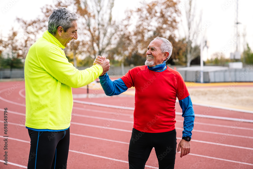 Two joyful senior men shaking hands on a track field, showcasing camaraderie and active lifestyle post-race. Two joyful senior men shaking hands on a track field, showcasing camaraderie and active lifestyle post-race.