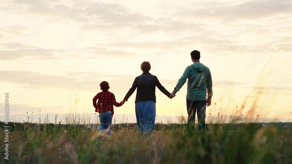 Happy family walks along grassy valley enjoying landscape at sunset. Sun rays fall on small family silhouettes walking along grassy valley. Family of parents and son walk along grass valley in evening