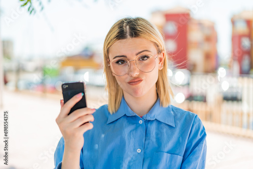Young pretty blonde woman using mobile phone at outdoors with sad expression Free Download Young pretty blonde woman using mobile phone at outdoors with sad expression - GFXLoad