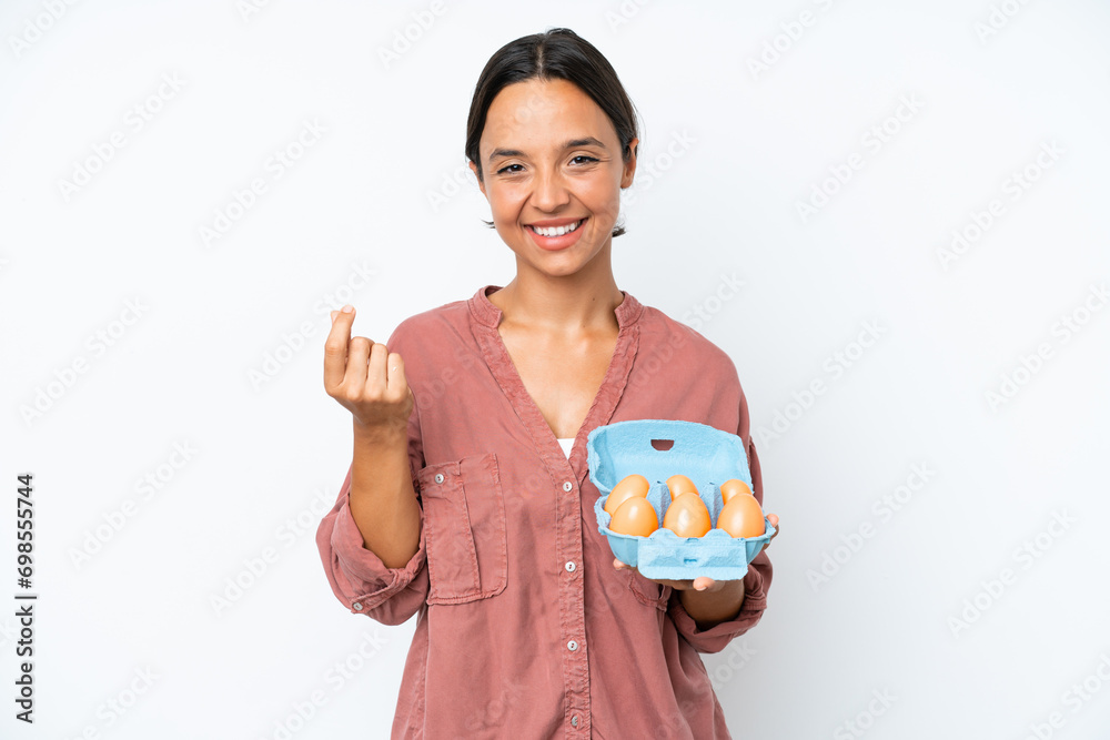 Young hispanic woman holding eggs isolated on white background making money gesture Young hispanic woman holding eggs isolated on white background making money gesture