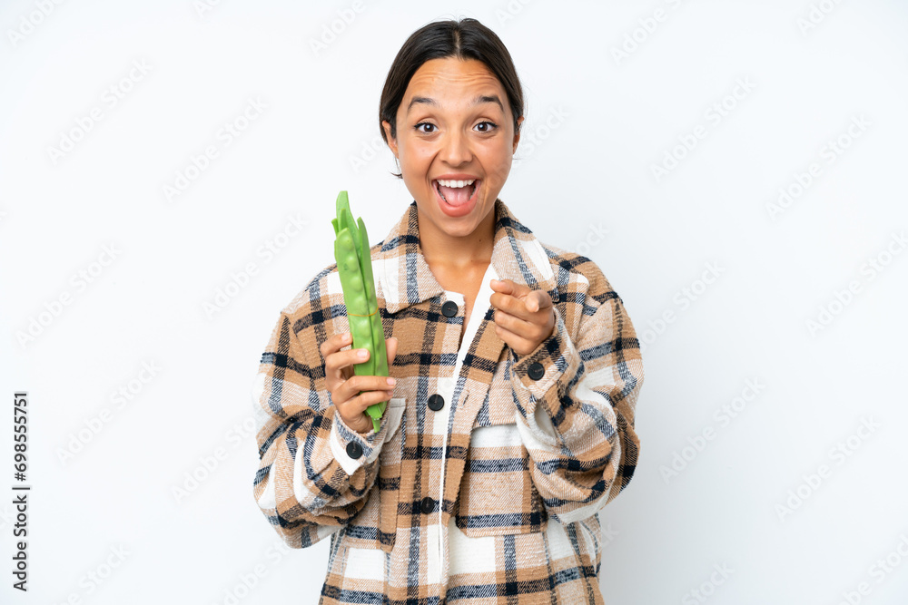 Young hispanic woman holding a green beans isolated on white background surprised and pointing front Young hispanic woman holding a green beans isolated on white background surprised and pointing front