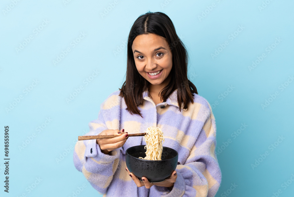 Young brunette mixed race woman over isolated blue background holding a bowl of noodles with chopsticks and offering it. Young brunette mixed race woman over isolated blue background holding a bowl of noodles with chopsticks and offering it.