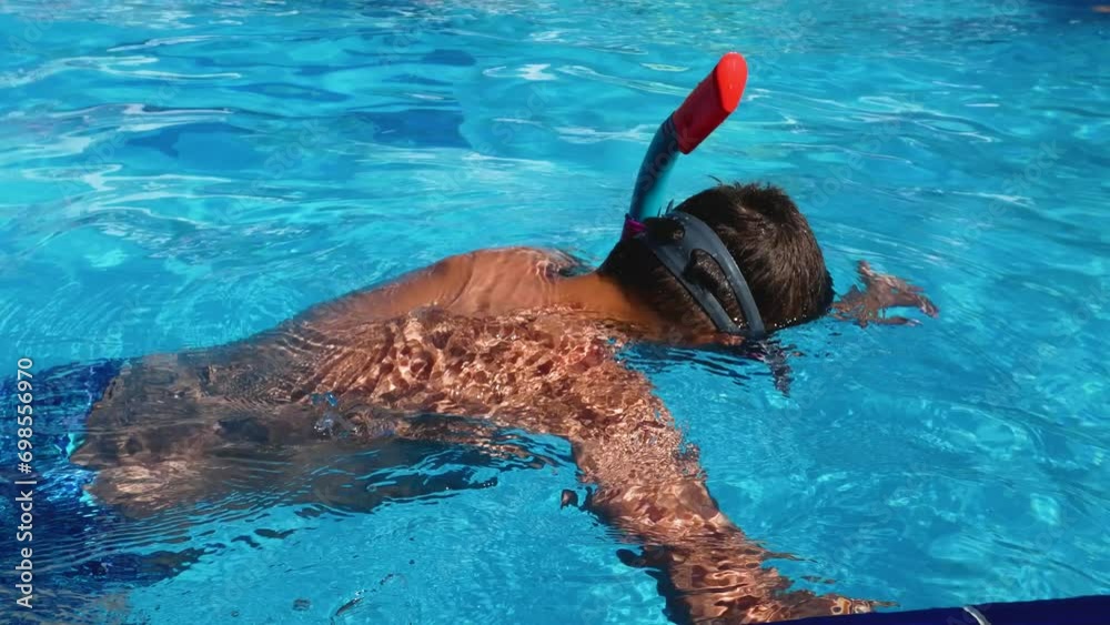 A boy in a swimming pool with a mask and snorkel holds on to the edge and dives into the pool. Summer fun by the pool. A child swims in the pool in summer with a mask and snorkel. Summer holidays