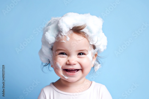 Little smiling baby girl with big soap foam on her head in her hair on a blue background