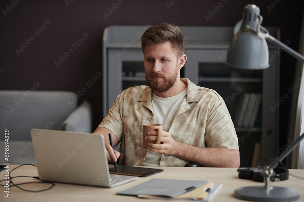 Portrait of young bearded man enjoying cup of coffee while working from home and using pen tablet for editing, copy space Portrait of young bearded man enjoying cup of coffee while working from home and using pen tablet for editing, copy space