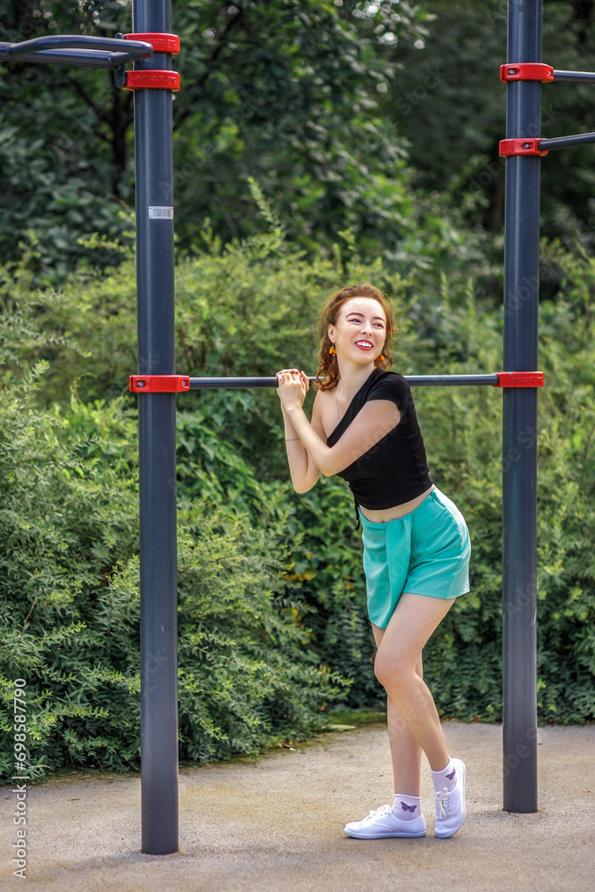A young girl in a sports bra and shorts leans against the horizontal bar on a sports field. Photo of a girl athlete with a beautiful athletic body