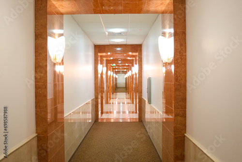 Long elegant hotel corridor white, beige and brown classical pattern on the walls. Clean, Sharp and straight hotel hallway. Interior design of walk way with wall lamp lights.