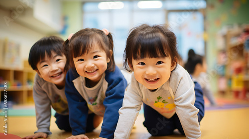 日本の幼稚園児・保育園児3人が長袖の私服で横に並んでハイハイでカメラに笑いながら向かってきている写真、背景教室、保育室のボケ photo
