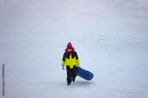 A little boy in warm bright outerwear carries a sled
