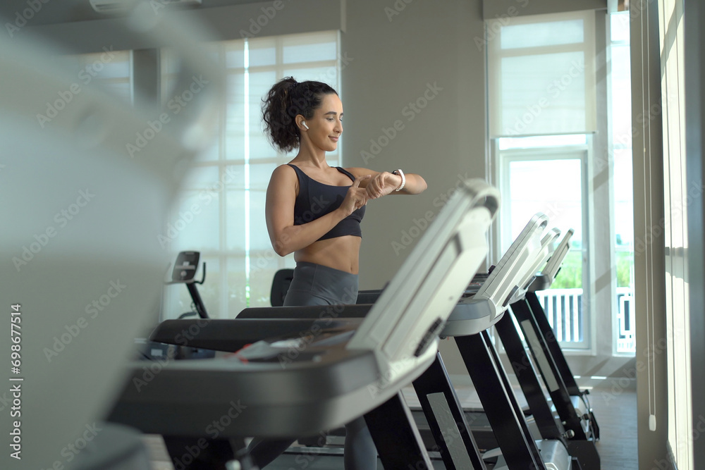Happy healthy hispanic woman practicing a cardio exercise by running and walking on treadmill machine in fitness and using fitness tracker or smartwatch to track workout progress.