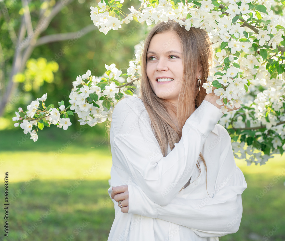 Face portrait of blonde young woman with cute smile wearing casual white blouse or shirt in the garden among blooming apple tree branches with small flowers and green leaves looking away at sunny day Face portrait of blonde young woman with cute smile wearing casual white blouse or shirt in the garden among blooming apple tree branches with small flowers and green leaves looking away at sunny day