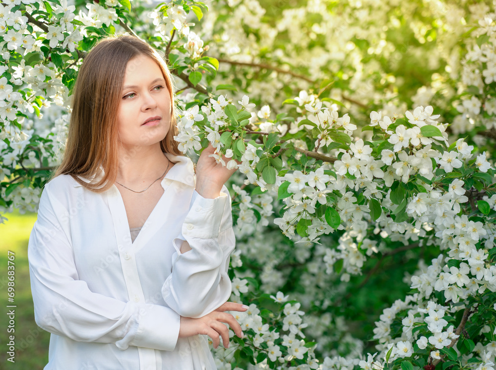 Portrait of blond young woman wearing casual white blouse or shirt in the garden among blooming apple trees with small flowers and green leaves standing with thoughtful smile at sunlight in spring Portrait of blond young woman wearing casual white blouse or shirt in the garden among blooming apple trees with small flowers and green leaves standing with thoughtful smile at sunlight in spring