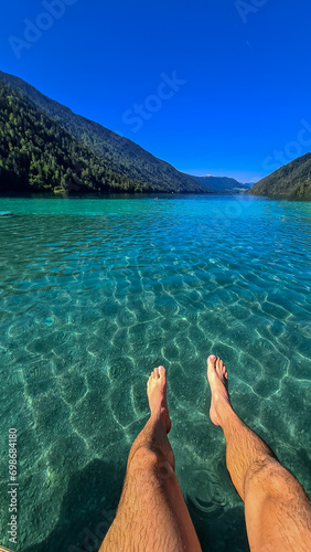 Close up on legs of man  lying at the east shore of Weissensee lake in Carinthia  Austria. Lake is surrounded by high Austrian Alps. Relaxation and chilling. Lake retreat in remote nature in summer