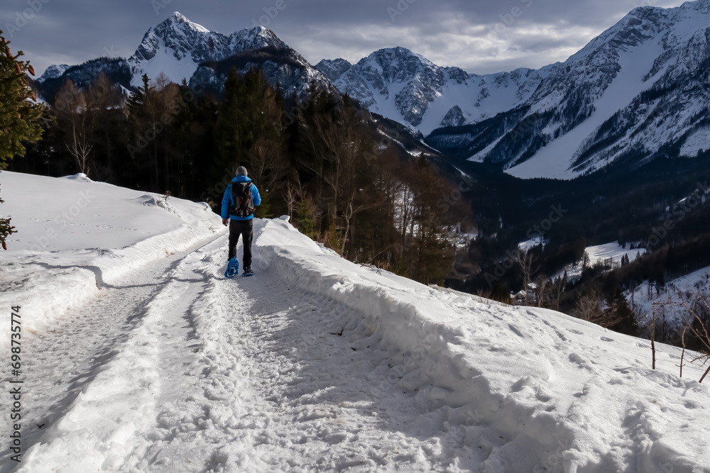 Man in snow shoes on hiking trail with panoramic view on snow capped mountain peaks of Karawanks, Carinthia, Austria. Looking at summits Kosiak and Hochstuhl, Austrian Alps. Alpine landscape in winter