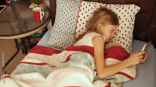 Sick little girl lying in bed covered with plaid watching her tablet