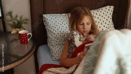 Cute little girl lying in bed watching cartoons on tablet looking at camera, sick child holding his toys in bed while feeling sick