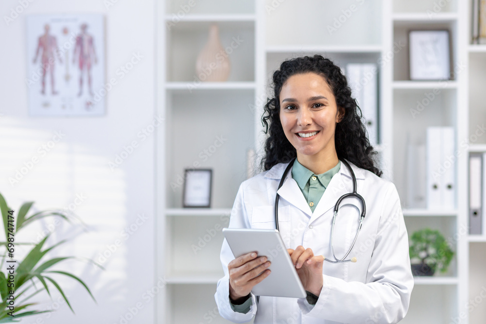 Close-up portrait of a young Latin American female doctor standing in the office in a white coat, holding a tablet and smiling at the camera. Close-up portrait of a young Latin American female doctor standing in the office in a white coat, holding a tablet and smiling at the camera.