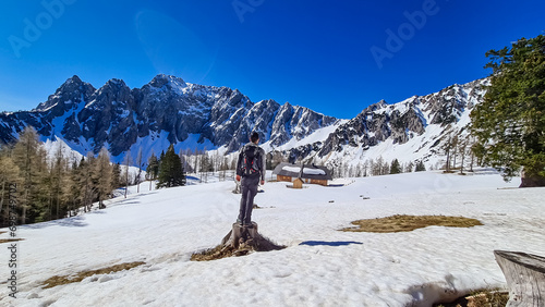 Hiker man standing on trunk on Ogrisalm enjoying scenic view of Karawanks mountains in Carinthia  Austria. Looking at snow capped summit of Vertatscha and Hochstuhl. Remote alpine cottage in Bodental