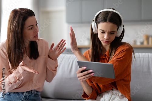 Teenage girl in headphones using digital tablet and gesturing stop to mother, while mom scolding daughter, sitting on sofa at home