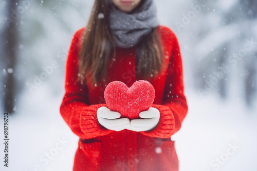 young woman holding a red woolen heart between her hands, wearing winter clothes, on snowing forest background, Valentine\'s Day concept