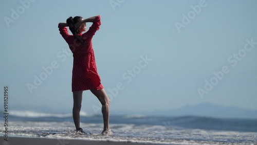 Serene woman standing ankle deep in water breaking seawave on sandy beach of ocean. Full length model generation x dressed in red polka dot babydoll dress loose fitting, hands behind head. Slow motion photo