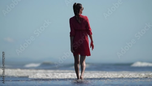 Back view woman ankle deep in sea water walking along beach on sunny day. Middle aged adult woman wearing red summer babydoll polka dot dress during beach holiday. Slow motion, cinematic handheld shot photo