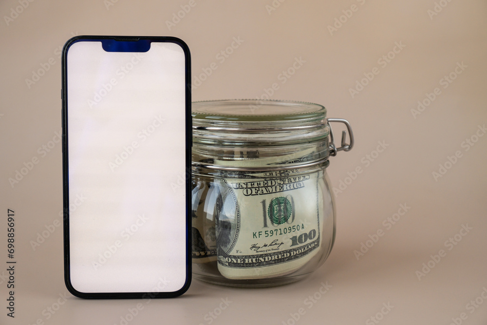 White smartphone screen mock up template in vertical position on beige background. Copy space App website advertising. Jar filled with dollars cash. Concept of Mobile application and technology White smartphone screen mock up template in vertical position on beige background. Copy space App website advertising. Jar filled with dollars cash. Concept of Mobile application and technology