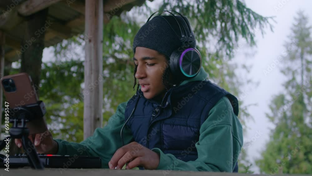 African American teenager wearing headphones plays on MIDI controller outdoors. Young musician, composer creates and records music while sitting in wooden gazebo, using phone on tripod. Slow motion. African American teenager wearing headphones plays on MIDI controller outdoors. Young musician, composer creates and records music while sitting in wooden gazebo, using phone on tripod. Slow motion.
