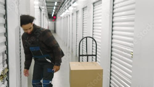 Young dilligent male loader in work overalls putting large cardpaper boxes into self storage unit with white metal roller doors in well lit long hallway. High quality 4k footage photo