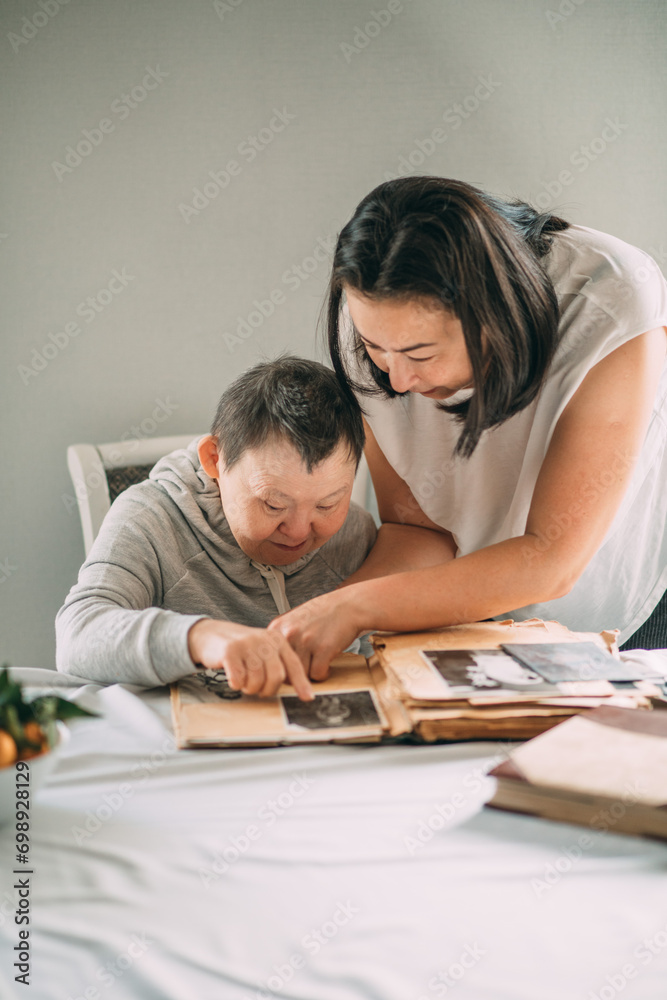 Asian girl points a smiling elderly woman with Down syndrome to a photo in an old album.