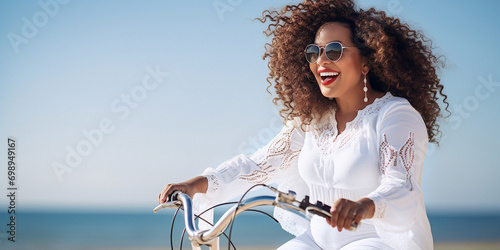 A young, plump, cheerful African-American hipster woman in a white suit rides a bicycle, in summer, along the seashore. Tropical vacation, summer trendy style, beach, cycling, travel, close-up, cheerf photo