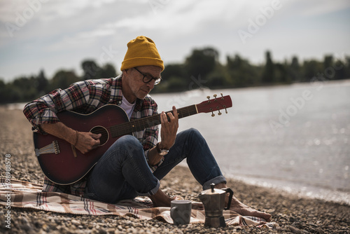 Senior man playing guitar sitting by sea on weekend photo