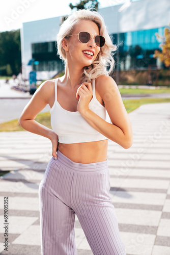 Young beautiful smiling hipster woman in trendy summer white top and violet pants clothes. Carefree model posing in the street at sunset. Positive blond female. Cheerful and happy. In sunglasses