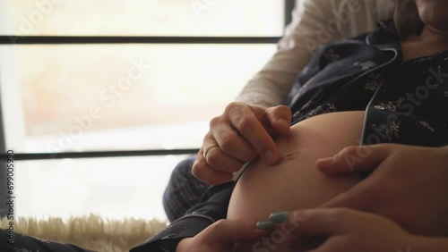 Faceless young pregnant woman in pajama relaxing with husband in bed at home. Close-up naked large belly touching by hands. Couple enjoying togetherness and waiting for a newborn baby