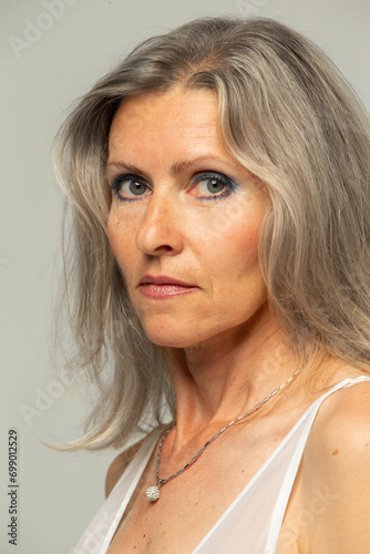 Beautiful grey-haired fifty year old woman looking at camera isolated on gray background. Mature old lady close up portrait. The face of an aging woman with wrinkles and imperfections.