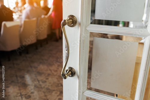 Close up of stylish stainless door knob or handle with metallic carvings on open white wooden modern interior door with glass. Concept of trends interior details.