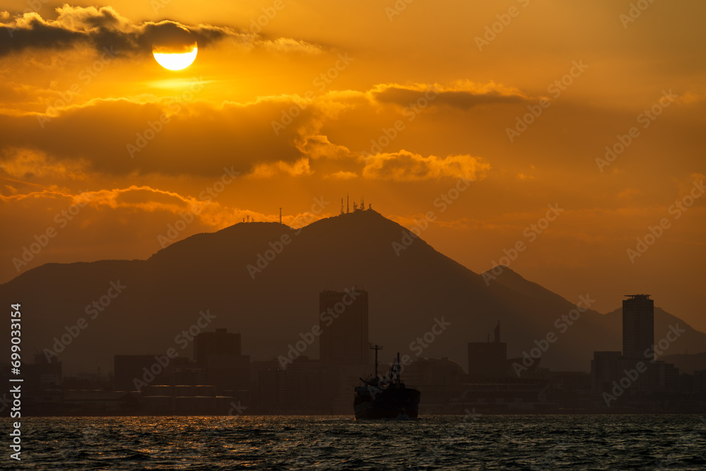 Fototapeta premium 関門海峡の夕日が照らす皿倉山と小倉の街影を背に船影を見る20231226