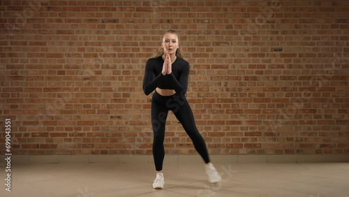 Fitness trainer performing Squat toe tap exercise. The girl is dressed in black sportswear and clearly demonstrates the physical exercise. She is against a brick wall in loft style. RED RAPTOR 8K RAW photo