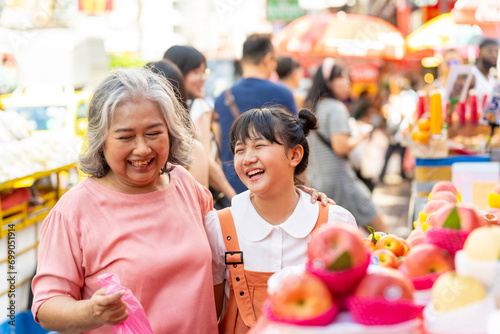 Happy Asian family grandmother and grandchild girl choosing and buying fresh fruit together at street market. Senior woman and little girl enjoy outdoor lifestyle travel in the city on summer vacation