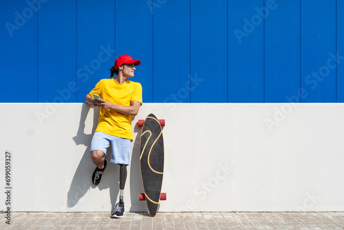 Young man with disability holding smart phone and standing near skateboard on sunny day photo
