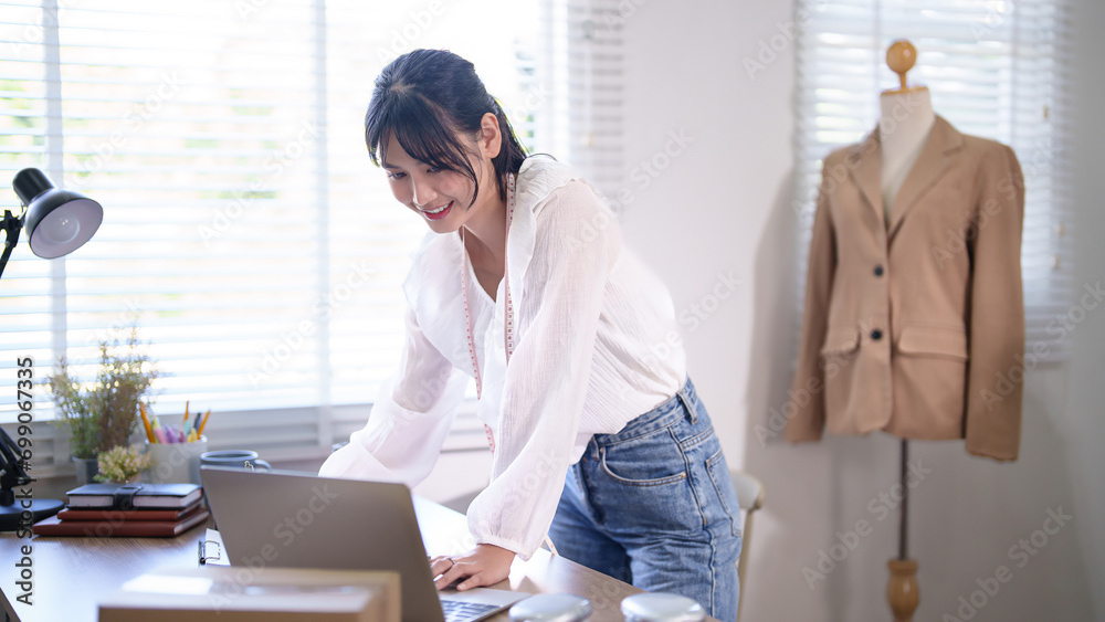 Obraz premium Young asian merchant women wearing measure tape in her neck and writing customer information in paper while checking purchase online order on laptop and working about online shopping business