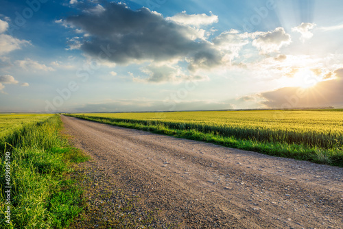 Country gravel road and green wheat field with sky clouds at sunset photo