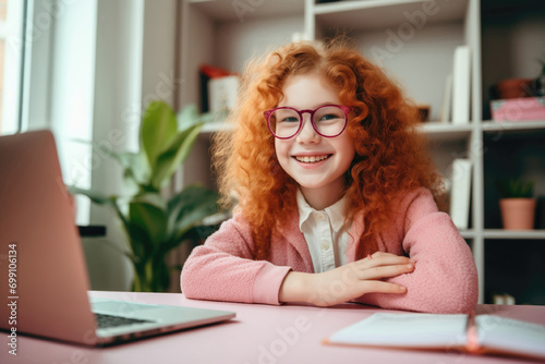 A girl with red curly hair wearing glasses sits at a desk behind a laptop and does lessons at home. Distance education, computer class.