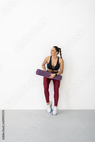 Smiling fitness woman standing in a fitness studio carrying a yoga mat.