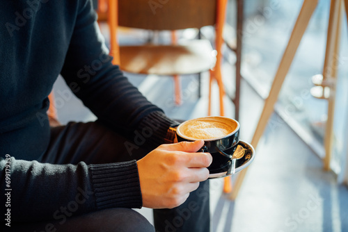 No face handsome young man in black clothes holding hot latte art or cappuccino coffee cup in modern cafe shop. Warm and cozy fall or winter moments. Take a break to relax. Soft selective focus photo