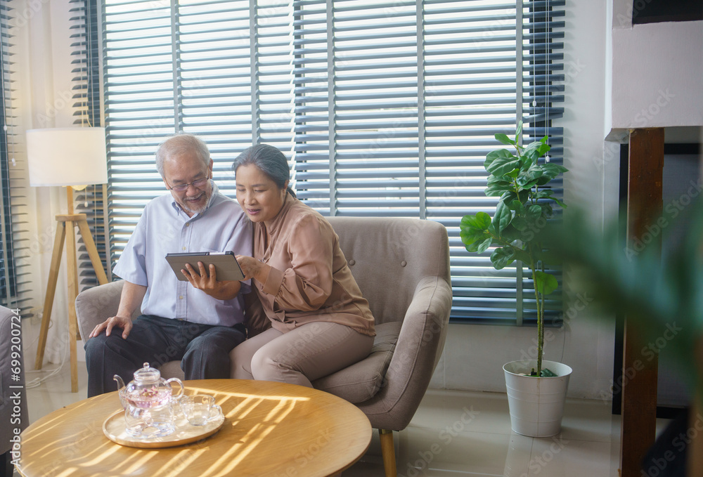 Asian grandfather and grandmother watching or meeting video call to family with digital tablet in the living room. Senior couple playing social media or searching on the internet. Asian grandfather and grandmother watching or meeting video call to family with digital tablet in the living room. Senior couple playing social media or searching on the internet.