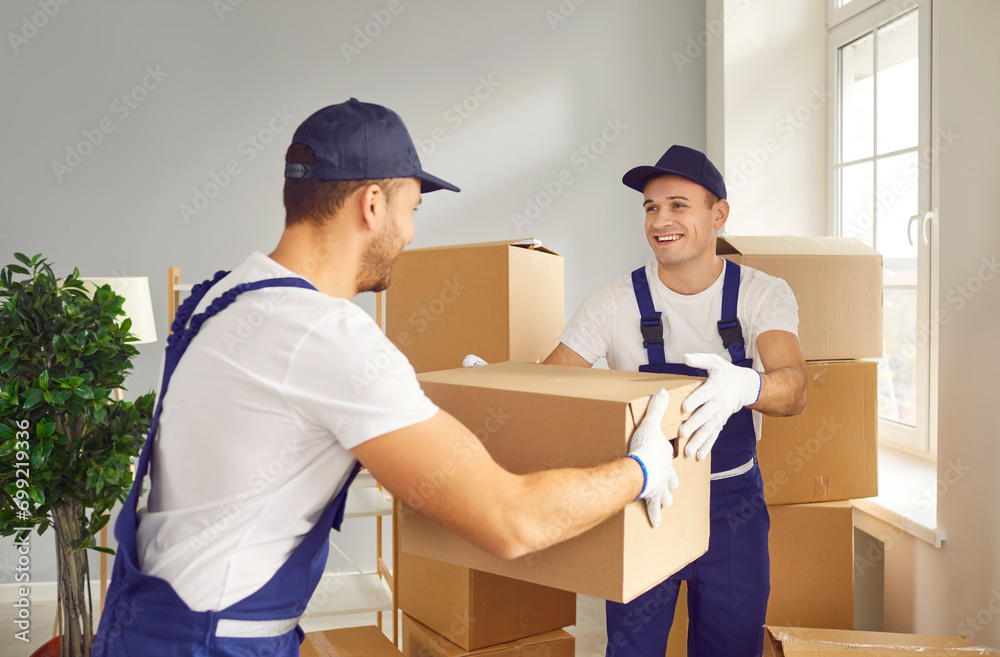 Team of two young happy cheerful professional workers of moving service in overalls carrying and delivering cardboard boxes in new apartment. Move, moving day and relocation concept. Team of two young happy cheerful professional workers of moving service in overalls carrying and delivering cardboard boxes in new apartment. Move, moving day and relocation concept.
